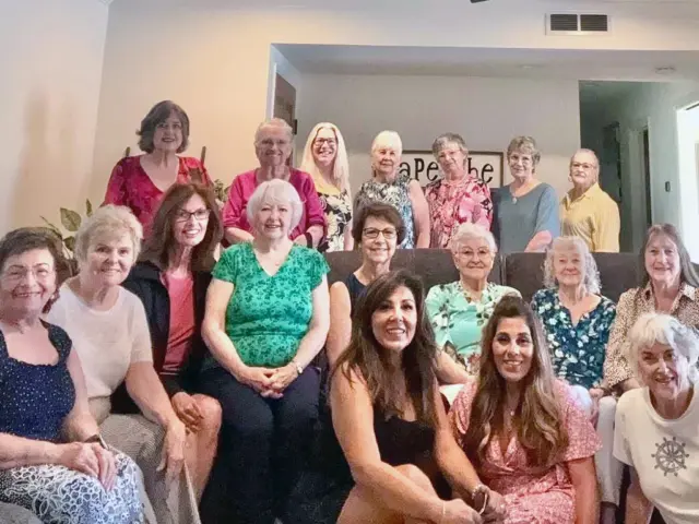 A group of women of various ages pose and smile together indoors, seated and standing in two rows in a living room.