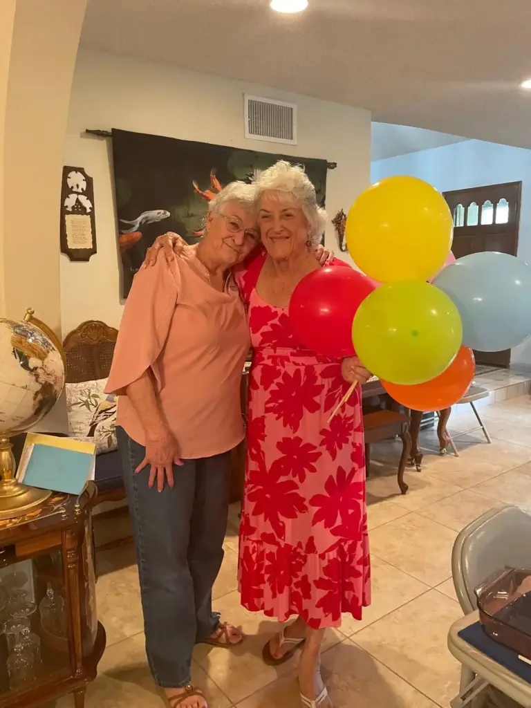 Two older women stand together indoors; one holds colorful balloons. they are smiling and appear to be celebrating an occasion in a living room setting.