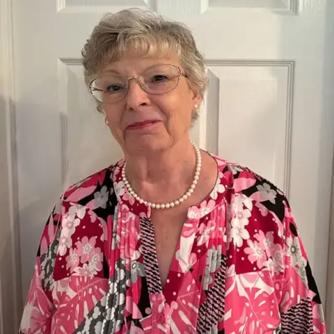 An older woman with short gray hair, glasses, and a pearl necklace stands in front of a white door, wearing a pink, black, and white floral blouse.