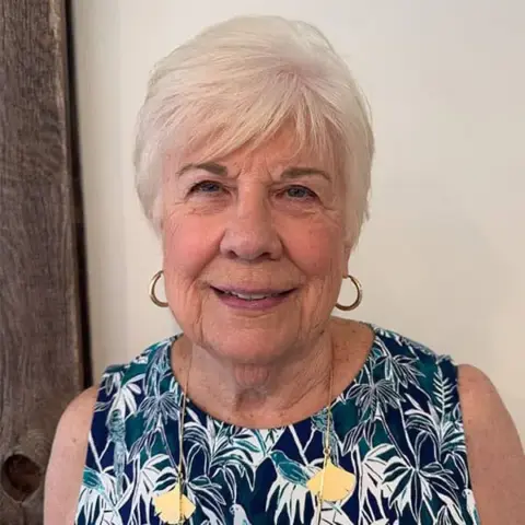 Older woman with short white hair, wearing gold hoop earrings and a blue floral sleeveless top, standing in front of a plain wall and wooden panel.