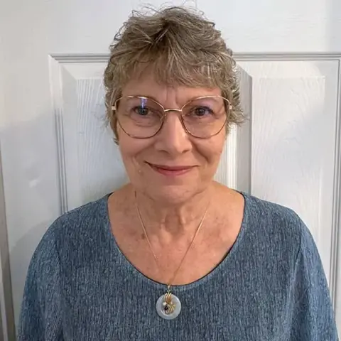 An older woman with short gray hair and glasses stands in front of a white door, wearing a blue top and a circular pendant necklace.