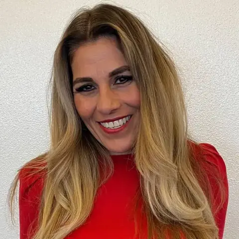 Woman with long, blonde hair wearing a red top, smiling in front of a plain, light colored background.