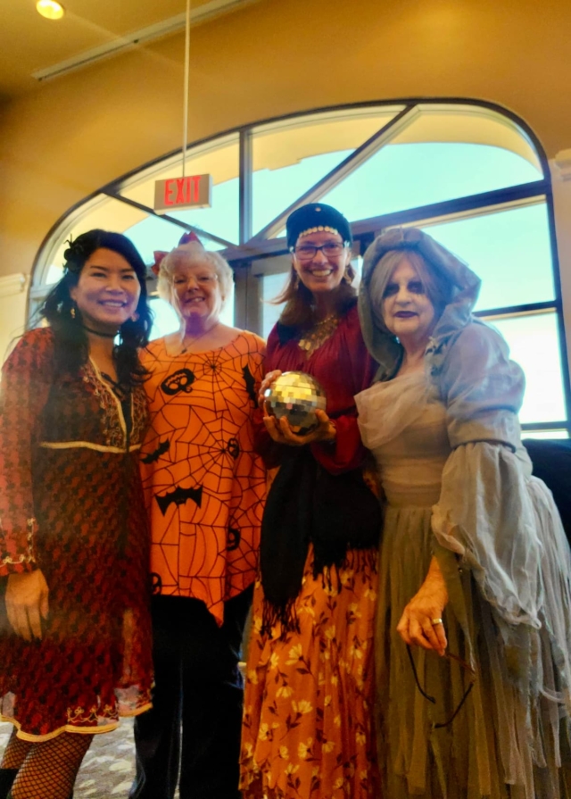 Four women wearing halloween costumes stand together indoors, smiling at the camera. one holds a shiny ball, and there is a large window in the background.