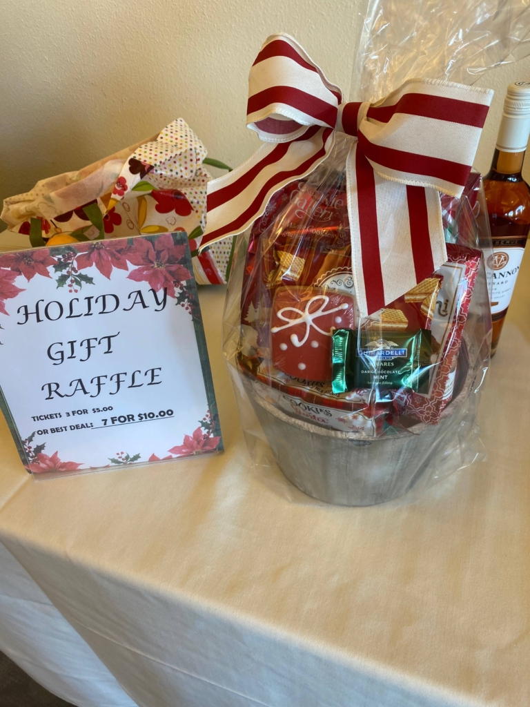 Gift basket wrapped in cellophane with a red and white bow next to a sign advertising a holiday gift raffle and a bottle of wine in the background.