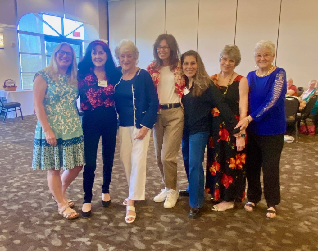 Seven women stand together and smile for a group photo in a large, well lit room with tables and chairs in the background.