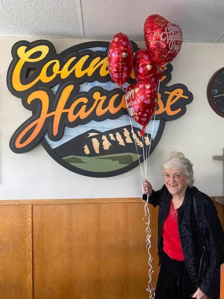 An older woman holds a bouquet of red heart shaped "Happy Valentine's Day" balloons in front of a "Country Harvest" sign inside a restaurant.