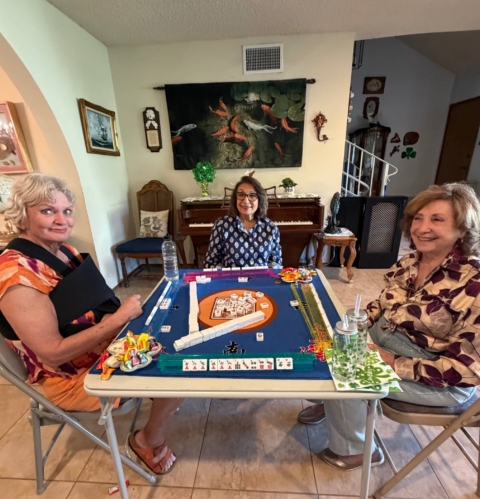 Three women sit around a table playing a game of mahjong in a living room, with game tiles, snacks, and drinks on the table.