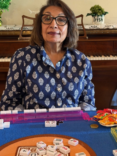 A woman with glasses sits at a table playing a tile game, with snacks and game pieces on the table and a piano in the background.