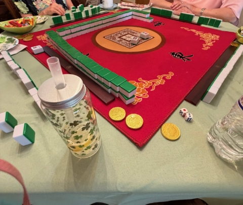 A group of people play mahjong on a red table mat; mahjong tiles, gold coin tokens, dice, and drink containers are visible on the table.
