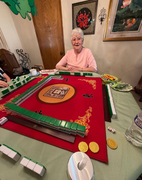 An elderly woman sits at a table playing mahjong on a red mat, surrounded by game tiles, dice, and snacks in a warmly decorated room.